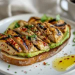 Grilled chicken slices served on avocado toast with a sprinkle of chili flakes and parsley, plated on a white ceramic plate.