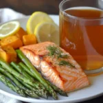 Baked salmon with asparagus and roasted sweet potatoes served on a white plate, garnished with lemon slices and fresh dill, accompanied by a glass of tea.