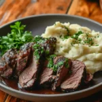 Sliced beef arm roast garnished with fresh parsley, served with creamy mashed potatoes on a dark plate, sitting on a rustic wooden table.