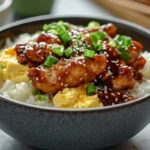 A bowl of oyakodon featuring tender chicken in a savory glaze, scrambled eggs, steamed white rice, garnished with sesame seeds and chopped green onions.